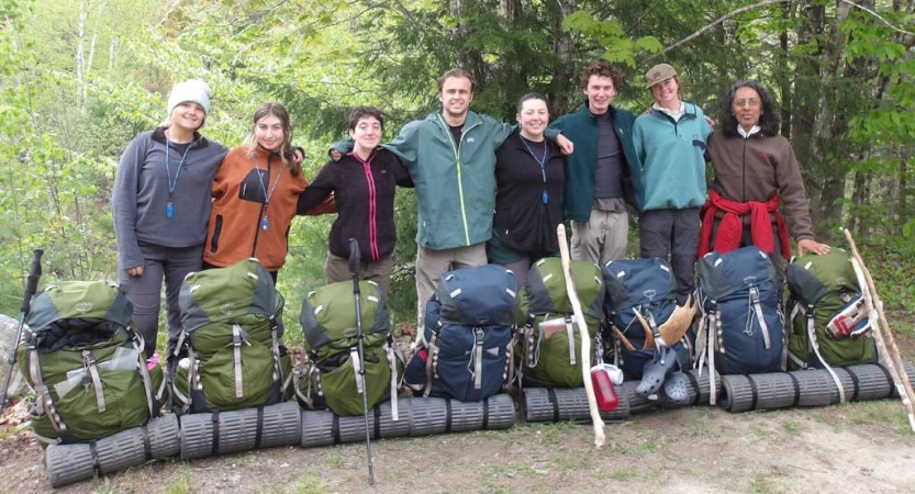 A group of people smile for a photo. Their backpacks rest on the ground in front of them.